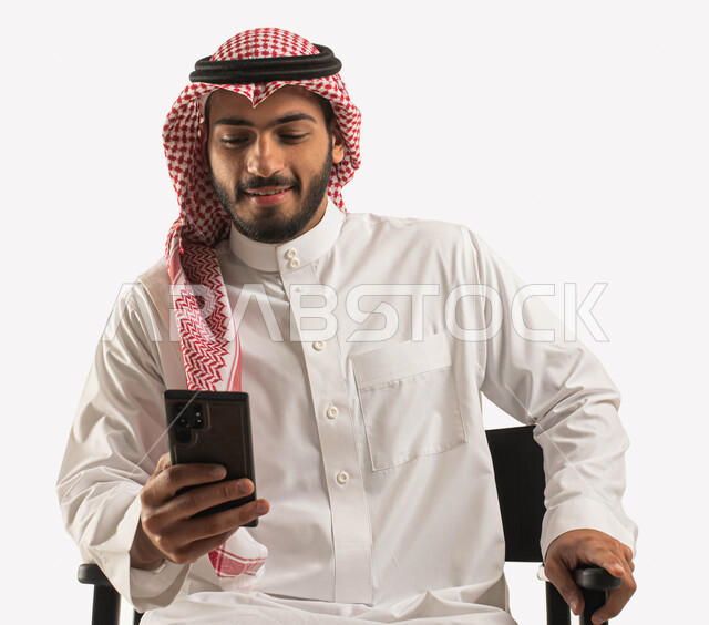 ايماءات الاندماج والتركيز، بورتريه مقرب لرجل عربي سعودي خليجي مبتسم يرتدي الشماغ و الثوب التقليدي يجلس على كرسي أسود ويستخدم الهاتف المحمول,، التحدث والدردشة مع الأصدقاء عبر الجوال، مفهوم الراحه والجلوس  , تصفح وسائل التواصل الاجتماعي، خلفية بيضاء