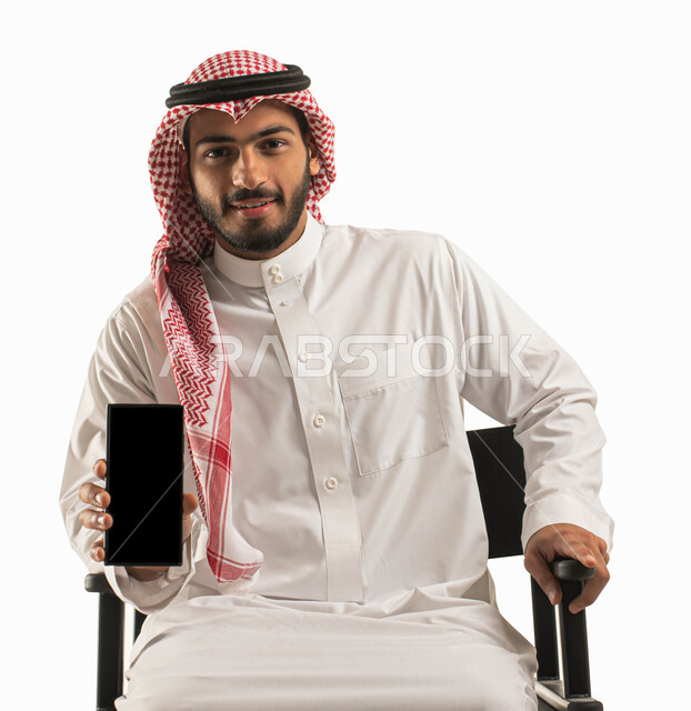 ايماءت الاعجاب باستخدام التقنيه والتكنولوجيا الحديثه، عرض تطبيقات وبرامج الهاتف المحمول، بورتريه لرجل عربي خليجي سعودي مبتسم يرتدي الشماغ والثوب التقليدي يحمل بيده جوال بشاشة سوداء فارغة، الراحه والجلوس على كرسي اسود،  مفهوم التسويق الالكتروني، خلفية بيضاء