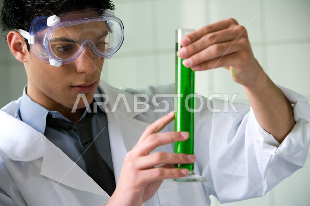 البيئة العملية المتطورة للقيام بالأنشطة التعليمة, البحث والتطوير لإجراء التفاعلات الكيميائية, صورة مقربة لطالب جامعي عربي خليجي إماراتي يقوم بتجربة علمية في المختبر, ارتداء نظارة الوقاية والمعطف الأبيض, اكتساب المعرفة والخبرة بإيماءات التركيز في التجربة