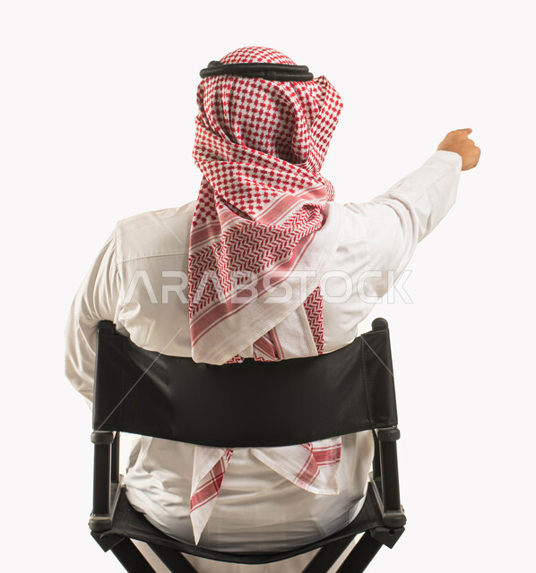 الإشارة إلى شيء أو مكان ما، بورتريه مقرّب من الخلف لرجل عربي خليجي سعودي يرتدي الثوب والشماغ التقليدي ويجلس على كرسي أسود مريح، مفهوم الجلوس والراحة، خلفية بيضاء