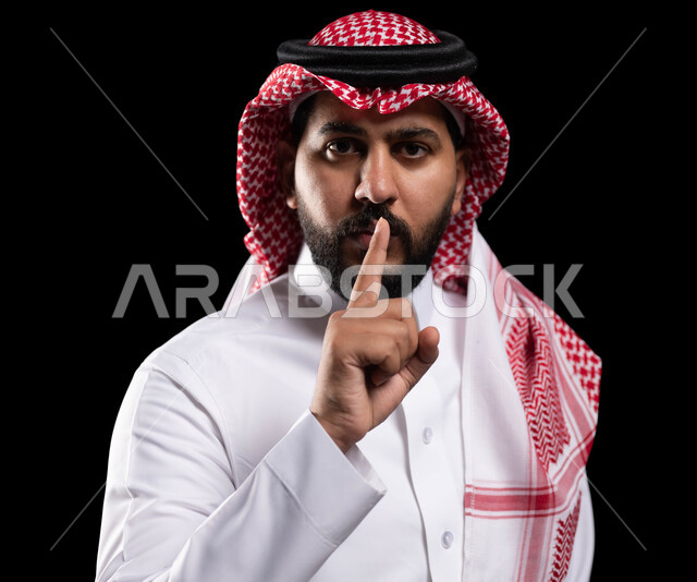 التوقف عن الكلام وخفض الصوت، بورتريه لمواطن عربي خليجي سعودي يرتدي الثوب التقليدي والشماغ ويضع أصبع السبابة على فمه، الهدوء والسكون وعدم الإزعاج، إيماءات وجه ويدين تدل على طلب السكوت والصمت، حركة أصبع وإشارات، خلفية سوداء