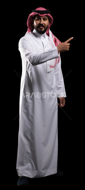 تعابير التحديد والتأكيد, الاشارة إلى شيء ما بإيماءات السرور ,بورتريه لرجل عربي خليجي سعودي مبتسم يرتدي الزي التقليدي والشماغ يشير بأصابع السبابة للاختيار وينظر إلى الكاميرا، صورة للجسم بالكامل، خلفية سوداء