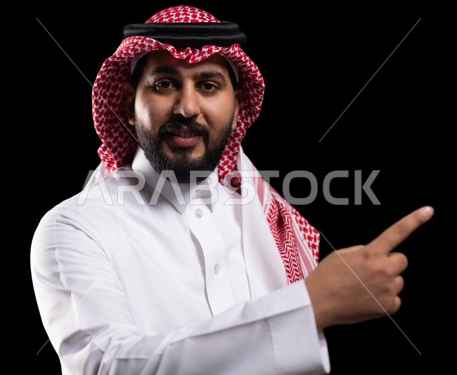 الاشارة إلى شيء ما بإيماءات السرور ,تعابير التأكد والتحديد, بورتريه مقرب لرجل عربي خليجي سعودي مبتسم يرتدي الزي التقليدي والشماغ يشير بأصابع السبابة للاختيار وينظر إلى الكاميرا، خلفية سوداء