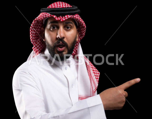 Pointing towards something and setting a goal, a close-up portrait of a Saudi Gulf Arab man wearing a shemagh and traditional dress and pointing with his index finger at a specific place with gestures of shock and astonishment, astonishment, astonishment and astonishment at something, black background.