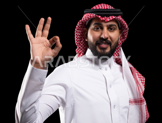 النظر إلى الكاميرا بإشارات الموافقه، حركات الاعجاب والقبول, بورتريه مقرب لرجل عربي خليجي سعودي مبتسم يرتدي الثوب والشماغ التقليدي يرفع يده للأعلى بإيماءات علامة الجودة و السرور, صورة الجسم بالطول الكامل، خلفية سوداء