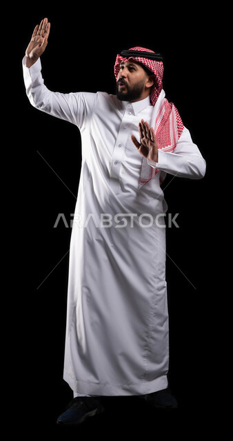 Standing cautiously and raising the hands with gestures of stopping and not approaching, caution, anxiety and staying away from something, a close-up portrait of a Saudi Gulf Arab man wearing the traditional thobe and shemagh protecting himself from external harm, full body portrait, black background