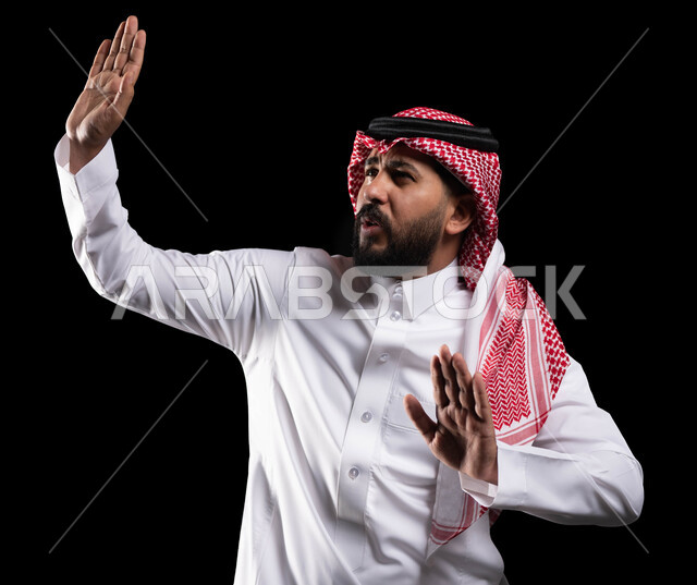 رفع اليدين بإيماءات التوقف وعدم الاقتراب ،الاحتياط والقلق والابتعاد عن شيء ما, حماية النفس من ضرر خارجي, بورتريه مقرب لرجل عربي خليجي سعودي يرتدي الثوب التقليدي والشماغ يقف بحذر, خلفية سوداء