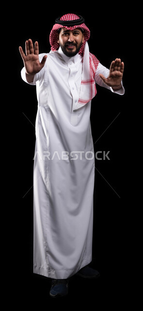 طلب التوقف والإمتناع عن فعل شيء ما، حماية النفس من ضرر خارجي, الرعب والذعر والرهبة من شيء ما، بورتريه لرجل عربي خليجي سعودي يرتدي الشماغ والزي التقليدي ويرفع كفيه لأعلى بإيماءات الخوف، صورة الجسم بالكامل، خلفية سوداء
