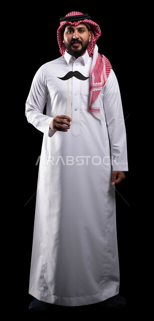 الاهتمام بصحة الرجل، الاحتفال بالمناسبات العالمية، بورتريه لمواطن عربي خليجي سعودي يرتدي الثوب التقليدي والشماغ ويمسك عود خشبي مع قصاصة ورقية على شكل شارب، اليوم العالمي للرجل 19 نوفمبر، صورة الجسم بالطول الكامل، خلفية سوداء