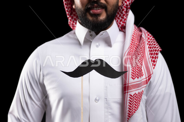 الاحتفال بالمناسبات العالمية، اليوم العالمي للرجل 19 نوفمبر، بورتريه مقرب لمواطن عربي خليجي سعودي يرتدي الثوب التقليدي والشماغ ويمسك عود خشبي مع قصاصة ورقية على شكل شارب، الاهتمام بصحة الرجل، خلفية سوداء