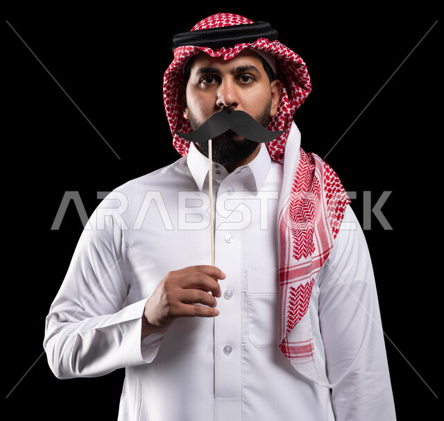 اليوم العالمي للرجل 19 نوفمبر، الاحتفال بالمناسبات العالمية، بورتريه مقرب لمواطن عربي خليجي سعودي يرتدي الثوب التقليدي والشماغ ويمسك عود خشبي مع قصاصة ورقية على شكل شارب، الاهتمام بصحة الرجل، خلفية سوداء