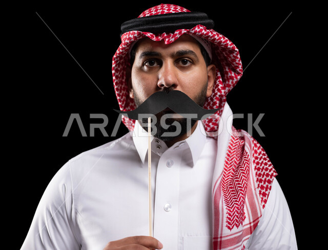 الاحتفال بالمناسبات العالمية، اليوم العالمي للرجل 19 نوفمبر، بورتريه مقرب لمواطن عربي خليجي سعودي يرتدي الثوب التقليدي والشماغ ويمسك عود خشبي مع قصاصة ورقية على شكل شارب، الاهتمام بصحة الرجل، خلفية سوداء