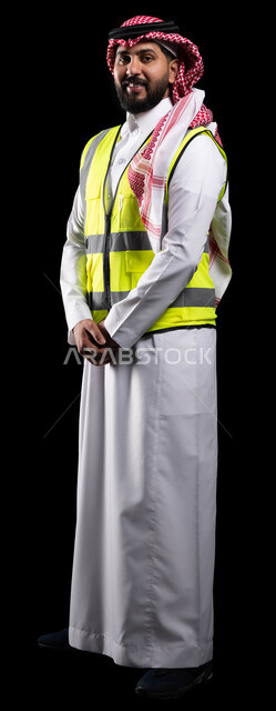 الوقوف باستقامة بإيماءات أيدي متماسكة، الإعمار بسواعد أبناء الوطن ،متابعة المشاريع الهندسية والرقابة عليها، بورتريه لمهندس مساعد عربي خليجي سعودي مبستم يرتدي الثوب التقليدي وسترة الحماية الخاصة بالعمل وينظر الى الكاميرا، صورة الجسم بالكامل، خلفية سوداء