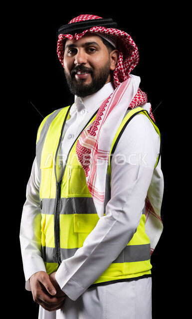 Reconstruction with the help of the people of the country, following up and monitoring engineering projects, a portrait of a smiling Saudi Arabian Gulf Arab assistant engineer wearing the traditional dress and work protection jacket and looking at the camera, standing straight with coherent hand gestures, engineering professions and jobs, black background
