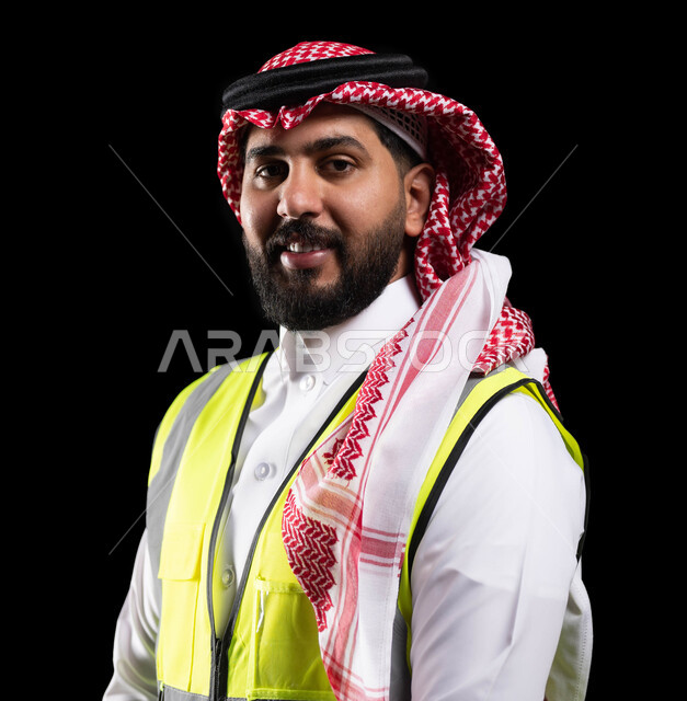 Supervision and control of engineering projects, portrait of a smiling Saudi Arabian Gulf Arab assistant engineer wearing the traditional dress and work protection jacket and looking at the camera, standing upright and confident, practicing the engineering profession in Saudi Arabia, black background