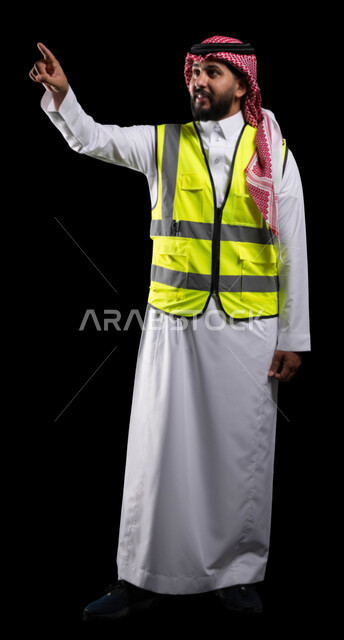 الاشارة باصبع السبابة باتجاه ما، بورتريه لمهندس معماري عربي خليجي سعودي يرتدي الشماغ والثوب التقليدي وسترة الحماية الخاصة بالعمل، النقر والضغط على عنصر ما، العمل في القطاع الهندسي السعودي، صورة الجسم بالطول الكامل، خلفية سوداء