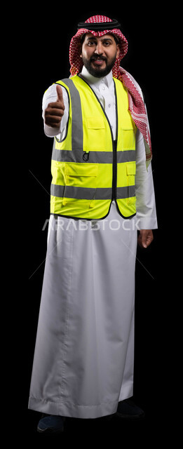 رفع اصبع الابهام للاعلى بايماءات القبول والموافقة والاعجاب، بورتريه لمهندس عربي خليجي سعودي يرتدي الزي التقليدي وسترة الحماية الخاصة بالعمل، متابعة المشاريع وتدقيق العمل، العمل في القطاع الهندسي السعودي، صورة الجسم بالطول الكامل، خلفية سوداء