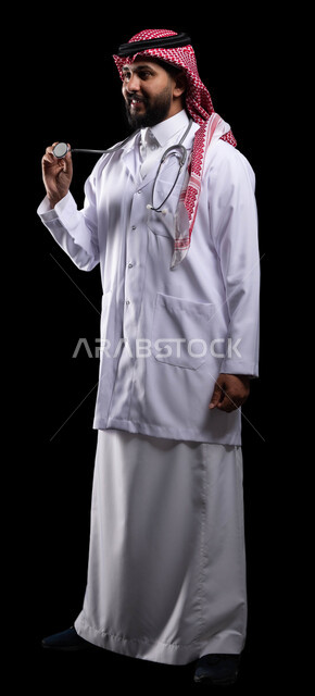 تأدية الخدمات والاستشارات الطبيه بحب واخلاص، وظائف ومهن سعودية ،إجراء الفحوصات اللازمة، بورتريه لطبيب عربي خليجي سعودي مبتسم يرتدي الزي التقليدي والمعطف الطبي، استخدام سماعة الطبيب لسماع نبضات القلب، صورة الجسم بالكامل، خلفية سوداء