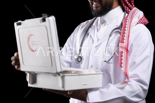 مفهوم الطب و تقديم الخدمات الصحية الطارئة للمرضى، التعامل مع الإصابات الشائعة وحالات الطوارئ، تفقد الأدوات والمعدات الطبية اللازمة، بورتريه مقرب لطبيب عربي سعودي خليجي يرتدي الثوب التقليدي و المعطف الطبي يحمل بيده حقيبة الاسعافات الأولية، خلفية سوداء