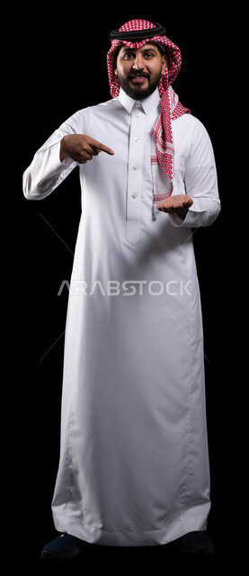 النظر إلى الكاميرا بإيماءات السرور، رفع وحمل شيء ما، بورتريه لرجل عربي خليجي سعودي مبتسم يرتدي الشماغ والثوب التقليدي يشير باصبع السبابة على عنصر ما، صورة الجسم بالطول الكامل، خلفية سوداء