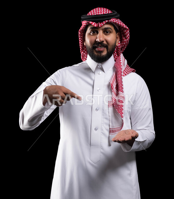 النظر إلى الكاميرا، ايماءات السعادة والسرور، رفع وحمل شيء ما، بورتريه لرجل عربي خليجي سعودي مبتسم يرتدي الشماغ والثوب التقليدي يشير باصبع السبابة على عنصر ما، خلفية سوداء