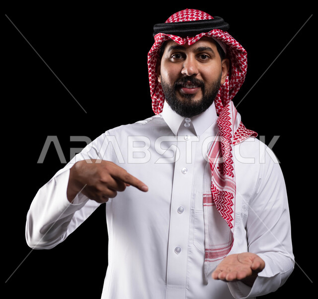 ايماءات السعادة والسرور، النظر إلى الكاميرا، رفع وحمل شيء ما،  بورتريه مقرب لرجل عربي خليجي سعودي مبتسم يرتدي الشماغ والثوب التقليدي يشير باصبع السبابة على عنصر ما، خلفية سوداء