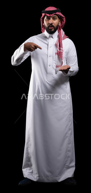 الاشارة باصبع السبابة على عنصر ما، حركات عين وفم تدل على الدهشة والاستغراب، النظر إلى الكاميرا، بورتريه لرجل عربي خليجي سعودي يرتدي الشماغ والثوب التقليدي متعجب من شيء ما، صورة الجسم بالطول الكامل، خلفية سوداء