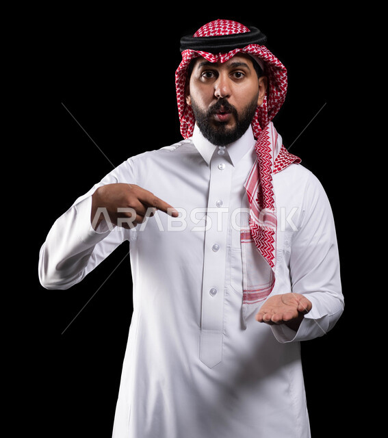 حركات عين وفم تدل على الدهشة والاستغراب، ايماءات الذهول والتعجب، النظر إلى الكاميرا، بورتريه لرجل عربي خليجي سعودي يرتدي الشماغ والثوب التقليدي ويشير بإصبع السبابة على عنصر ما، خلفية سوداء