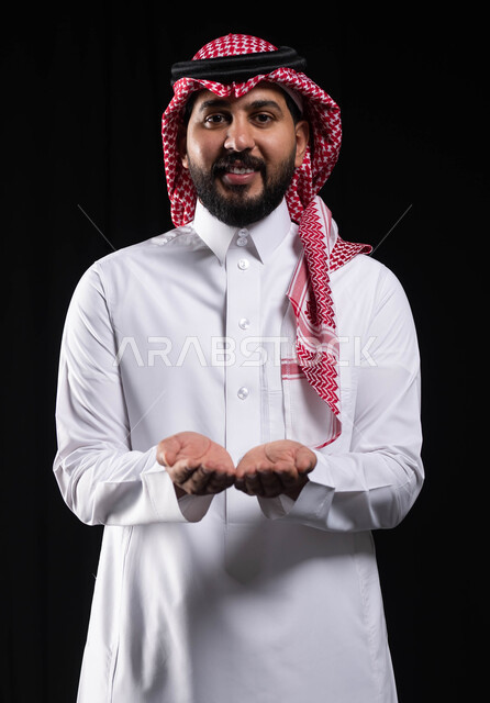 النظر إلى الكاميرا بإيماءات السرور والبشاشة, بورتريه لرجل عربي خليجي سعودي مبتسم يرتدي الشماغ والثوب التقليدي يفتح يديه لعرض شيء ما، حمل ورفع عنصر ما على كفيه، خلفية سوداء