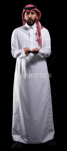 ايماءات الدهشة والذهول، بورتريه لرجل عربي خليجي سعودي يرتدي الشماغ والثوب التقليدي يفتح يديه لعرض شيء ما، حمل ورفع عنصر ما على كفيه، الوقوف باستقامة والنظر إلى الكاميرا, صورة الجسم بالطول الكامل، خلفية سوداء