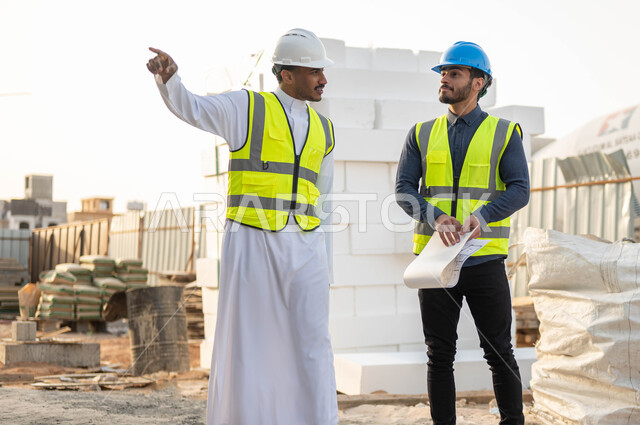النقاش والحوار وتبادل الأفكار, التحقق من تطبيق الشروط الصحيحه للبناء, الإشاره والنظر إلى شيء ما, مهندسان عربيان سعوديان خليجيان يرتديان الستره وخوذه الحمايه يتجولان في موقع العمل, الاهتمام بالأساسيات الجيده للمبنى, متابعه سير الخطة التنظيميه للمشروع