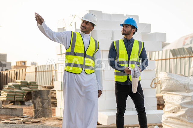 التحقق من تطبيق الشروط الصحيحه للبناء, الإشاره والنظر إلى شيء ما, مهندسان عربيان سعوديان خليجيان يرتديان الستره وخوذه الحمايه يقفان في موقع العمل, الاهتمام بالأساسيات الجيده للمبنى, متابعه سير الخطة التنظيميه للمشروع, النقاش والحوار وتبادل الأفكار