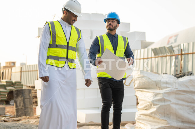 التشاور وتبادل الأفكار بإيماءات السرور, مهندسان عربيان سعوديان خليجيان مبتسمان يرتديان الخوذه وستره الحماية يقفان في موقع العمل, متابعه سير الخطة التنظيميه للمشروع, التحقق من تطبيق الخطط  المعمارية في المبنى, المخططات والتصاميم الهندسيه للمنشآت السعوديه