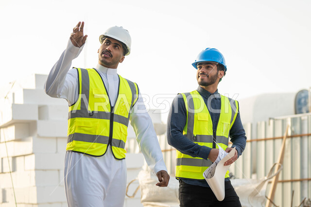 Verifying the application of the correct construction conditions, pointing and looking at something, two Saudi and Gulf Arab engineers wearing jackets and protection helmets standing at the work site, paying attention to the good foundations of the building, following up on the progress of the project’s organizational plan, discussion, dialogue and exchange of ideas.
