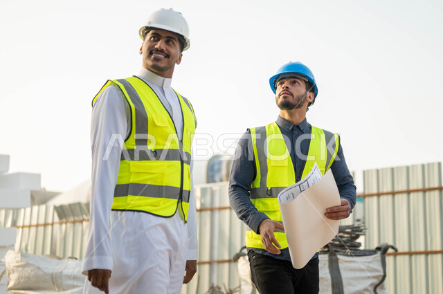 المخططات والتصاميم الهندسيه للمنشآت السعوديه, مهندسان عربيان سعوديان خليجيان مبتسمان يرتديان الخوذه وستره الحماية ينظران إلى شيء ما بإيماءات السرور, متابعه سير الخطة التنظيميه للمشروع, التحقق من تطبيق الخطط  المعمارية في المبنى