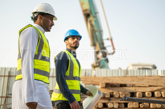 Supervising and walking on the construction site, working in the engineering sector, two Saudi Gulf Arab engineers and contractors wearing protective jackets and helmets checking the implementation of the building’s architectural plans, looking at something with gestures of interest.