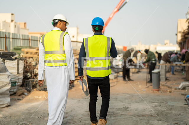 Touring and supervising the construction site, working in the engineering sector, a picture from the back of an Arab engineer and contractor, two Gulf Arabs, two Saudis wearing a protective jacket and helmet, verifying the application of the architectural engineering plans for the building, a group of workers preparing steel iron.