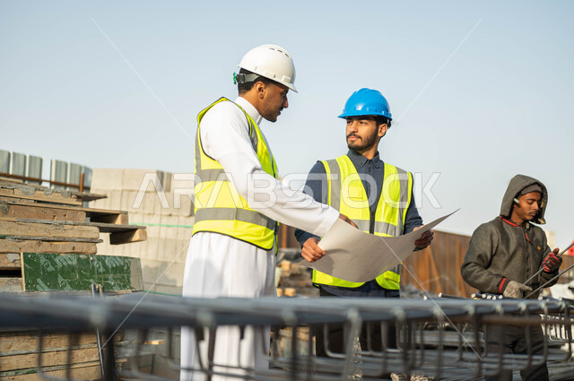 Following up on the progress of the organizational chart for architectural projects, construction and engineering designs in Saudi Arabia, paying attention to the good foundations of the building, two Saudi and Gulf Arab engineers wearing helmets and protective vests standing at the work site, exchanging ideas about completing the engineering project.