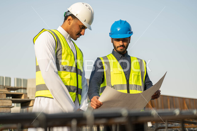 الاهتمام بالأساسيات الجيده للمبنى, مهندسان عربيان سعوديان خليجيان يرتديان الخوذه وستره الحمايه يقفان في موقع العمل, تبادل الأفكار حول إنجاز المشروع الهندسي, متابعه سير المخطط التنظيمي للمشاريع المعماريه, الإنشاءات والتصاميم الهندسيه في السعوديه
