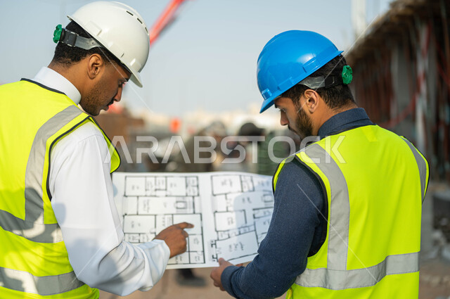 Following up and auditing the progress of architectural projects, working in the engineering field, standing at the construction site, dialogue and discussion about the construction project, a close-up photo from the back of a Saudi-Gulf Arab contractor and engineer wearing helmets and protection vests, holding the construction drawings plan.