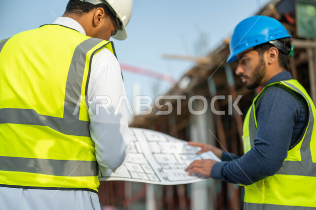 Auditing and following up on the progress of architectural projects, working in the engineering field, standing at the construction site, dialogue and discussion about the construction project, a close-up photo from the back of a Saudi-Gulf Arab contractor and engineer wearing helmets and protection vests, holding the construction drawings plan.