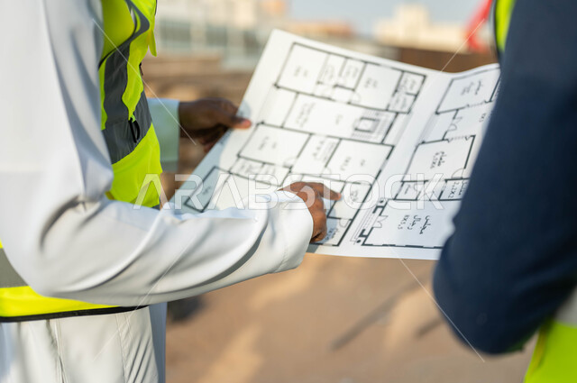 الإشاره إلى الرسومات والمخططات الإنشائية, صورة مقربة لمهندسيين عربيين خليجيين سعوديين يمسكان مخطط المشروع, الوقوف في موقع العمل, الإشراف والتدقيق على خطة البناء, الاهتمام بالأساسيات الجيده للمبنى
