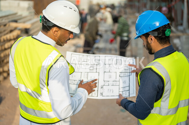 الحوار والنقاش حول مشروع البناء, صورة مقربه من الخلف لمقاول ومهندس عربيين سعوديين خليجيين يرتديان الخوذه وستره الحمايه يمسكان مخطط الرسومات الإنشائيه, التدقيق ومتابعه سير المشاريع المعماريه, العمل في المجال الهندسي, الوقوف في موقع الإنشاء