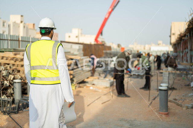 مفهوم الهندسه والإنشاءات, صورة من الخلف لمهندس عربي خليجي سعودي يرتدي الخوذه وستره الحمايه يمسك مخطط البناء, الإشراف على سير خطة المشروع, العمل في المجال الهندسي, مجموعه من العمال يقومون بإنجاز المهام  المعماريه