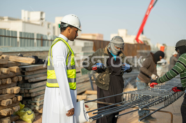 Developing the construction sector and the concept of engineering and construction, a Saudi Arabian Gulf engineer wearing a helmet and a protective vest holding a construction plan, supervising the progress of the project plan, working in the engineering field, a group of workers completing architectural tasks