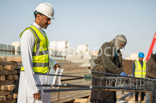 العمل في المجال الهندسي, مجموعه من العمال يقومون بتجهيز الحديد الصلب, مهندس عربي خليجي سعودي مبتسم يرتدي الخوذه وستره الحمايه يمسك مخطط الإنشاءات في موقع البناء, الاشراف على تطبيق المشاريع والخطط الهندسيه, المهاره والدقه في إنجاز المهام