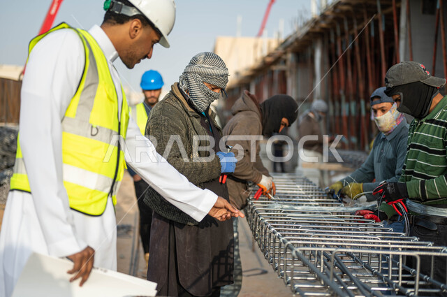 النمو والتطور العمراني بسواعد أبناء الوطن, المهاره في اتقان العمل, مهن ووظائف سعوديه, مجموعه من العمال يقومون بتجهيز وقص الحديد الصلب بمعدات وأدوات البناء, مهندس عربي خليجي سعودي يرتدي الستره وخوذه الحمايه يقوم بالإشراف ومتابعه سير االمشاريع المعماريه