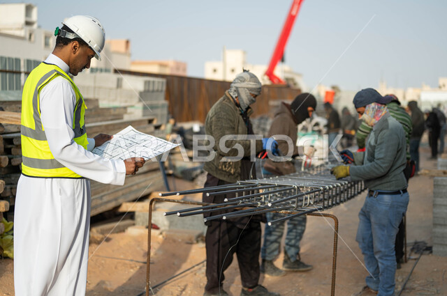 مهن ووظائف سعوديه, مجموعه من العمال يقومون بتجهيز وقص الحديد الصلب بمعدات وأدوات البناء, مهندس عربي خليجي سعودي يرتدي الستره وخوذه الحمايه ينظر إلى مخطط الرسومات والإنشاءات الهندسيه, الدقه والمهاره في اتقان العمل