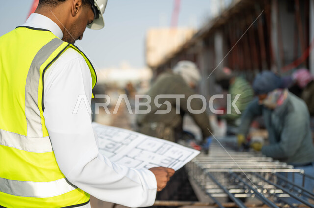 الرسومات والمخططات الإنشائيه, الطموح في العمل لتحقيق النجاح, صورة مقربه من الخلف لمهندس عربي سعودي خليجي يرتدي ستره الحمايه والخوذه ينظر إلى البناء, الاهتمام في جوده المواد الأوليه, التحقق من تطبيق المشاريع والخطط الهندسيه
