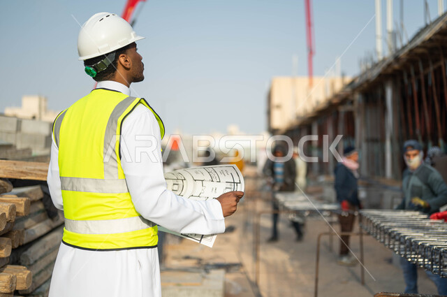 المخططات والرسومات الإنشائيه, الطموح في العمل لتحقيق النجاح, صورة من الخلف لمهندس عربي سعودي خليجي يرتدي ستره الحمايه والخوذه ينظر إلى البناء, الاهتمام في جوده المواد الأوليه, التحقق من تطبيق المشاريع والخطط الهندسيه